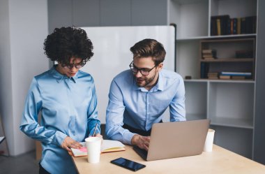 İş adamı dizüstü bilgisayar kullanıyor ve kız arkadaşının not defterine ne yazdığına bakıyor. İş birliği ve takım çalışması. Masadaki genç Kafkas milenyumu insanları. Modern başarılı insanlar