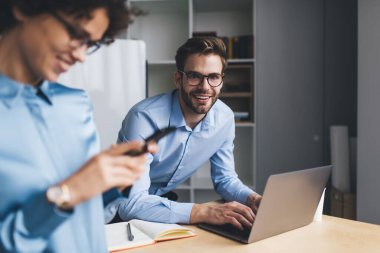 Masada dizüstü bilgisayarda çalışan gülümseyen iş adamı ve bayan meslektaşı cep telefonu kullanırken kameraya bakıyor. Modern başarılı insanlar kavramı. Genç Avrupalı milenyum meslektaşları görevde