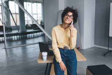 Milenyum Kafkasyalı iş kadını akıllı telefondan konuşuyor ve kameraya bakıyor. Modern başarılı kadın kavramı. Açık uzay ofisindeki tahta masada gözlüklü, gülümseyen kıvırcık kız.