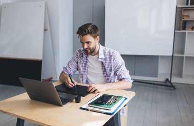 Milenyum Kafkas işadamı laptoptan izliyor ve grafik tabletine bir şeyler yazıyor. Modern başarılı adam kavramı. Açık uzay ofisindeki masada oturan genç odaklı sakallı adam.