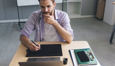 Bin yıllık beyaz iş adamı laptoptan izliyor ve grafik tabletine bir şeyler yazıyor. Modern başarılı adam kavramı. Açık uzay ofisindeki masada oturan genç sakallı adam.