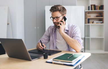 Milenyum Kafkas işadamı akıllı telefondan konuşuyor, dizüstü bilgisayardan izliyor ve grafik tabletine yazı yazıyor. Modern başarılı adam kavramı. Açık uzay ofisindeki masada oturan gözlüklü genç adam.