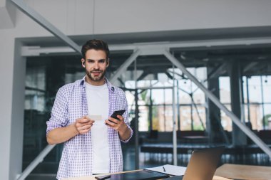 Milenyum Kafkas işadamı elinde kartvizit ve akıllı telefon tutarken kameraya bakıyor. Modern başarılı adam kavramı. Açık uzay ofisindeki masada kablosuz kulaklıklı genç adam.