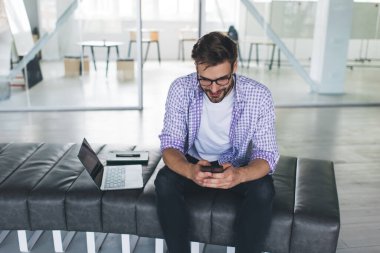 Milenyum Kafkas işadamı akıllı telefondan daktilo ediyor ve izliyor. Modern başarılı adam kavramı. Deri bankta oturan gözlüklü yakışıklı ve açık uzay ofisinde dizüstü bilgisayarı olan genç adam.