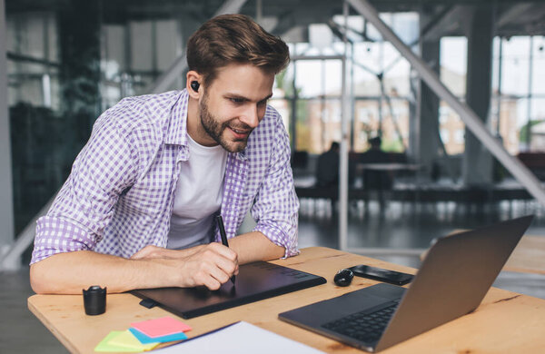 Millennial caucasian businessman watching on laptop and writing something in graphics tablet. Concept of modern successful man. Young smiling guy in wireless earphones at desk in open space office