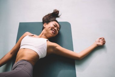 Omurga bükme pozisyonunda yoga yapan Asyalı bir kadın. Uyum ve akıl sağlığı kavramı. Genç, gülümseyen, gözleri kapalı spor giysili spor ayakkabılı ve spor minderinde çıplak ayaklı bir kız.