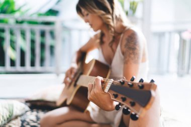Otelin terasında akustik gitar çalan genç bir kadının bulanık görüntüsü. Eğlence, eğlence ve hobi anlayışı. Tatil ve hafta sonu fikri. Halıda oturan dövmeli kız.