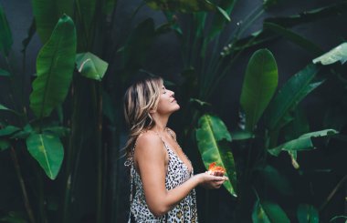 Tropikal bahçede gözleri kapalı, çiçekli, hayalperest genç bir kadın. Turizm, tatil ve hafta sonu. Elbise giyen çekici beyaz bir kız. Bali adasında huzurlu ve huzurlu bir yaşam tarzı