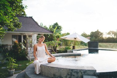 Otelin bahçesinde havuz kenarında oturup dinlenen gülümseyen genç bir kadın. Turizm, tatil ve hafta sonu. Güzel beyaz bir kız. Bali adasında huzurlu ve huzurlu bir yaşam tarzı. Güneşli bir gün