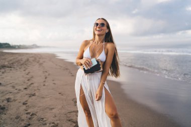 Elektronik kulaklıklı neşeli bayan milenyum edebiyat kitabı tutuyor ve deniz kıyılarında eğlenmek için boş zamanlarında gülüyor, güneş gözlüklü neşeli kadın pozitif mobil müzik dinliyor.