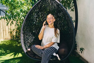 Klasik gözlüklü mutlu beyaz kız uluslararası mobil sohbetin tadını çıkarırken avluda sallanan sallanan sandalyede dinleniyor, gözlüklü neşeli milenyum uygulaması aracılığıyla arıyor.