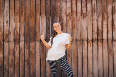 Neşeli bir mankenin yarım boy portresi, ahşap promosyon arka planının yakınındaki olumlu fotoğraf çekimi sırasında, kameraya gülümseyen, optik gözlüklü mutlu bir Kafkas genci gösteriyor.