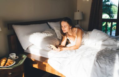 Kafkas kadın blogcu rahat bir yatakta cep telefonuyla ağlardaki web sayfalarına göz atmak için yatıyordu. Hippi kız, otel odası servisini kullanarak mobil cihazdan sabah uyarısı alıyordu.