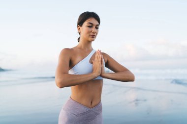 Eşofmanlı kadın yogi Hatha namaste pozunda meditasyon yapıyor. Kıyı şeridinde rahatlama ve konsantrasyon zamanının tadını çıkarıyor.