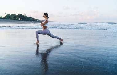 Eşofmanlı konsantre bir kadın bacak egzersizi yapıyor zayıflama için ilham alıyor, sabah deniz kıyısındaki antrenmanda yoga yapmaktan zevk alıyor.