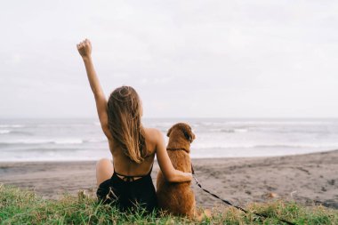 Kıyı şeridindeki boş zamanlarında en sevdiği köpeğe sarılan beyaz bir kadının arkası. Gezgin kız, sevimli bir hayvanla birlikte hafta sonunun keyfini çıkarıyor.
