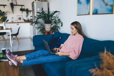 Avrupalı bir kız evdeki kanepede yatarken dizüstü bilgisayar kullanıyor. Ev hayatı kavramı. Ev istirahatı ve boş zaman fikri. Kazak, kot pantolon ve spor ayakkabı giyen genç bir kadın. Stüdyo dairenin içi.