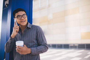 Asyalı milenyum çocuğu akıllı telefondan konuşur ve şehirdeki otobüs durağına bakar. Uzaktan kumanda ve serbest çalışma kavramı. Modern başarılı adam. Kahveli, gülümseyen genç bir erkek. Güneşli bir gün
