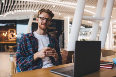 Masada akıllı telefon kullanırken kameraya bakan beyaz bir genç. Çevrimiçi eğitim ve iş kavramı. Modern, başarılı bir adamın fikri. Ofiste çalışan ya da ders çalışan gözlüklü gülen adam.