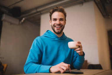 Kafeteryadaki boş zamanlarında elinde bir fincan sıcak çay tutan ve kameraya gülümseyen mutlu hipsterin yarım boy portresi. Kafeteryada kahve içip eğlenen neşeli beyaz adam.