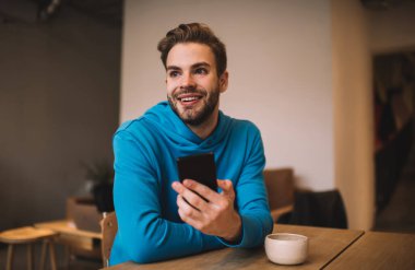 Hücresel teknolojiye sahip mutlu beyaz erkek kafeterya içinde eğleniyor, neşeli milenyum adamı dijital akıllı telefon aleti tutuyor ve kahve dükkanında boş zamanlarında gülümsüyor.