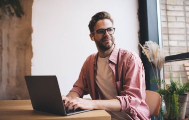 Kafenin iç kısımlarında uzaktan müzik dinleyen beyaz bilişim uzmanı, modern dizüstü bilgisayarda bluetooth 'a bağlı elektronik kulaklıklar takan yetenekli bir erkek.