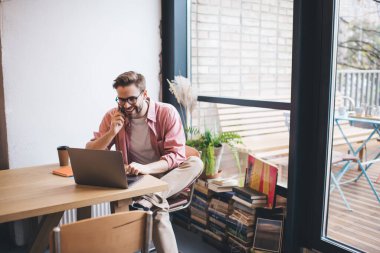 Optik gözlüklü neşeli erkek serbest çalışan cep telefonuyla konuşan mutlu hipster çocuk çevrimiçi kurs öğrenirken cep bilgisayarında araştırma yapıyor.