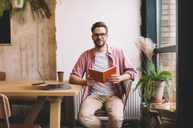 Optik gözlüklü genç bir erkek öğrencinin portresi, elinde eğitim defteri, çalışma alanında öğrenme, 20 'li yaşlarda, ders kitabı ve dizüstü bilgisayarı olan beyaz bir adam kameraya bakıyor.