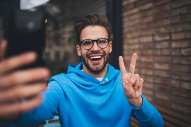 Sosyal medyaya paylaşmak için video içeriği çekerken cep telefonu kamerasına gülümseyen mutlu milenyum kullanıcısı, akıllı telefonuyla medya uygulaması üzerinden selfie çekerken V işareti gösteren neşeli erkek blogcu