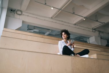 Gülümseyen bin yıllık beyaz bir kızın akıllı telefonuyla bakışlarının alttaki görüntüsü. Mola ve iş yerinde dinlenme kavramı. Genç kıvırcık kadın ahşap bankta oturuyor.