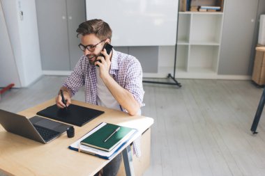 Milenyum Kafkas işadamı akıllı telefondan konuşuyor, dizüstü bilgisayardan izliyor ve grafik tabletine yazı yazıyor. Modern başarılı adam kavramı. Ofisteki masada oturan gözlüklü genç adam.