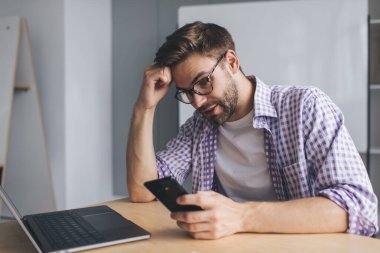 Bin yıllık beyaz iş adamının iş sırasında akıllı telefondan bir şey izlemesine şaşırdım. Modern başarılı adam kavramı. Gözlüklü genç, açık uzay ofisinde dizüstü bilgisayarla masada oturuyor.