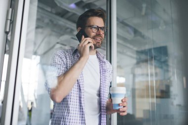 Milenyum Kafkas işadamı akıllı telefondan konuşuyor ve başka tarafa bakıyor. Modern başarılı adam kavramı. Gözlüklü, kahve fincanı olan, açık uzay ofisinde duran genç, sakallı adam.