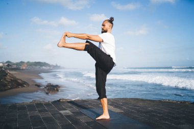 Spor kıyafetleri içinde beyaz bir erkek spor yaparken vücut kaslarını esnetiyor siyah paspas üzerinde sabahları kıyı şeridinde esnek bir adam açık havada bütünsel iyileşme için geri çekiliyor.