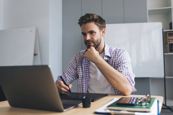 Millennial caucasian businessman watching on laptop and writing something in graphics tablet. Concept of modern successful man. Young concentrated guy sitting at table in open space office