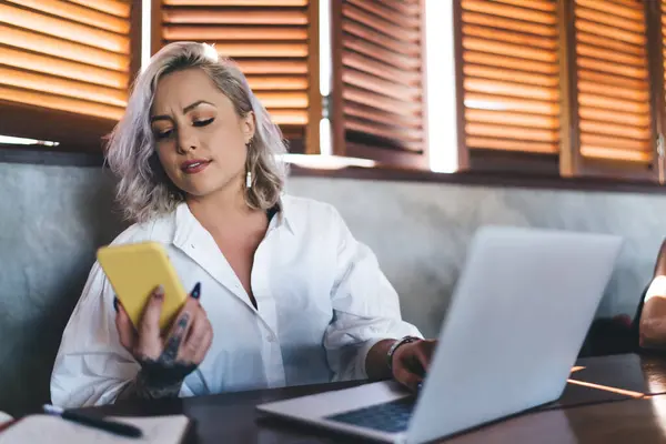 Stock image Caucasian copywriter sharing text content using bluetooth connection on modern cellphone and laptop technology, puzzled female IT professional checking received notification on mobile device