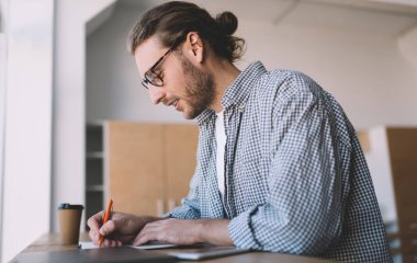 Optik göz giysisi giyen zeki bir erkek öğrencinin ödev yaparken eğitim bilgisi yazması, gözlüklü zeki bir hipsterın üniversite sınavlarına hazırlanırken not alması.