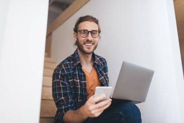 Klasik gözlüklü neşeli erkek programcının portresi dizüstü bilgisayar ve akıllı telefon aygıtı kullanarak uzaktan iş yaparken, cep telefonu ve netbook teknolojisiyle kameraya gülümseyen mutlu adam.