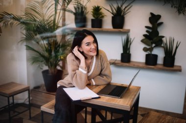 Kafedeki dizüstü bilgisayarda çalışırken kameraya bakan Asyalı bir girişimci. Genç gülümseyen milenyum esmer kız masada oturuyor. Serbest çalışma ve uzak çalışma kavramı. Modern kadın yaşam tarzı