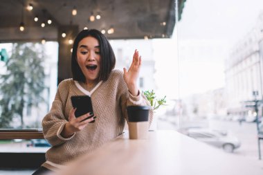 Akıllı telefondan izleyen ve kafede el kol hareketi yapan Asyalı bir kız. Genç milenyum esmer kadın pencerenin yanındaki masada duruyor. Dinlenmek, boş zaman ve boş zaman kavramı. Modern kadın yaşam tarzı