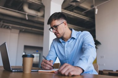 Optik gözlüklü beyaz erkek öğrenci, eğitim ortamında öğrenim görürken internetten eğitim ağı seyrediyor, gözlük konusunda yetenekli, dizüstü bilgisayarla uzak mesafe çalışması yaparken grafik tasarımı yapıyor.