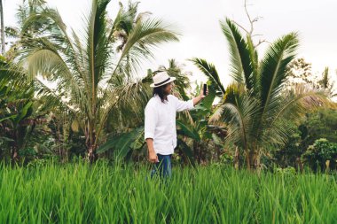 Ubud 'daki pirinç tarlalarında yürürken 4G kablosuz cihazıyla selfie çekmeyi seven şapkalı mutlu erkek turist Bali' de internetten iletişim çağrısında bulunuyor.