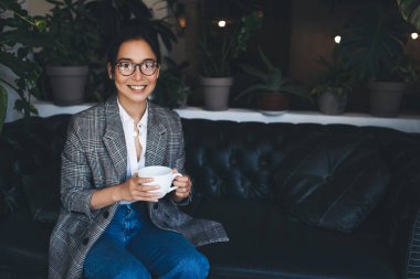 Asyalı başarılı bir modelin elinde bir fincan sıcak çay tutarken ve içerde eğlenmek için kameraya gülümserken portresi görme düzeltmesi için optik gözlüklü mutlu bir kadın.
