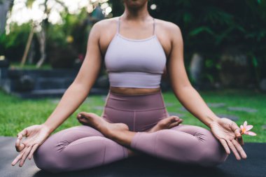 Lotus 'ta yoga yapan bir kızın dış görünüşü. Uyum ve akıl sağlığı kavramı. Genç atletik kadın spor giysisi giyer spor minderine oturur ve Bali adasının bahçesinde egzotik çiçekleri tutar.