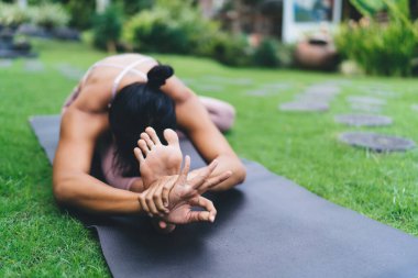 Yoga yapan bir kız, baş-diz öne kıvrılıp dışarıda poz veriyor. Uyum ve akıl sağlığı kavramı. Genç esmer atletik kadın spor giysisi giyiyor Bali adasının bahçesinde jimnastik minderinde yatıyor.