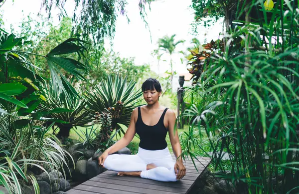 Lotus 'ta yoga yapan Asyalı kız dışarıda poz veriyor. Uyum ve akıl sağlığı. Gözleri kapalı genç atletik kadın spor giyer ve tropik bahçedeki tahta tahtalara çıplak ayakla oturur. Bali adası