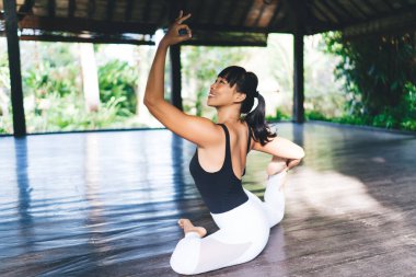 Gülen Asyalı kız tek bacaklı kral güvercin pozuyla yoga yapıyor. Sağlıklı yaşam tarzı kavramı. Tahta terasta spor giysisi giyen genç ve güzel bir bayan. Bali adasında güneşli bir gün