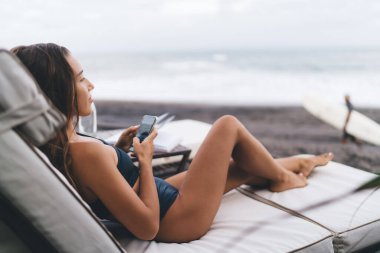 Genç seyahat blogcusu dinlenme odasında dinleniyor. İnternet ağı kurmak için modern mobil teknolojiyi kullanıyor. Mayolu kadın, güneşlenirken cep telefonu cihazında 4G kablosuz internete bağlanıyor.