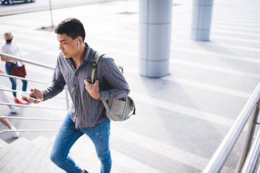Genç Asyalı milenyum çocuğu akıllı telefon kullanıyor ve merdivenlerden dışarı çıkıyor. Uzaktan kumanda ve serbest çalışma kavramı. Modern, başarılı bir adamın fikri. Çantalı kulaklıklı bir erkek. Güneşli bir gün