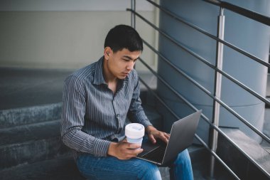 Asyalı milenyum çocuğu ofis binasının yanındaki merdivenlerde otururken dizüstü bilgisayardan izliyor. Serbest çalışma ve uzak çalışma kavramı. Modern, başarılı bir adamın fikri. Kahveli genç bir erkek. Güneşli bir gün